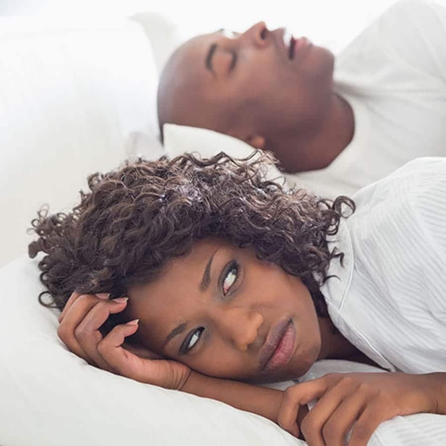 annoyed woman lays in bed awake as her husband snores next to her.