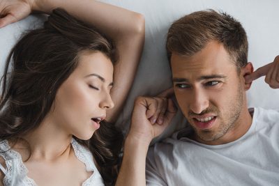 man plugs his ears while woman snores loudly next to him